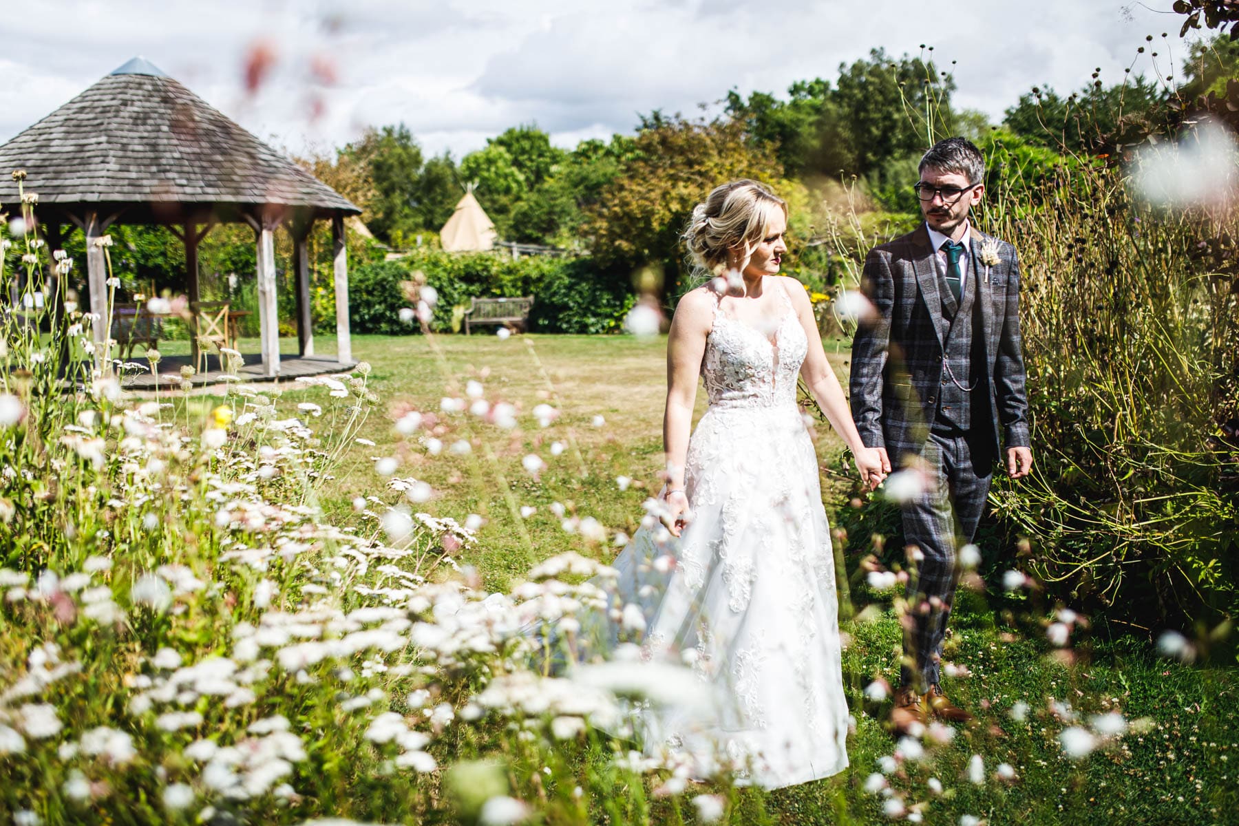 A bride in a white dress and a groom in a checked suit walk hand-in-hand through a garden filled with white wildflowers at The Gardens wedding venue, with a wooden gazebo and green trees captured by the Kent wedding photographer.
