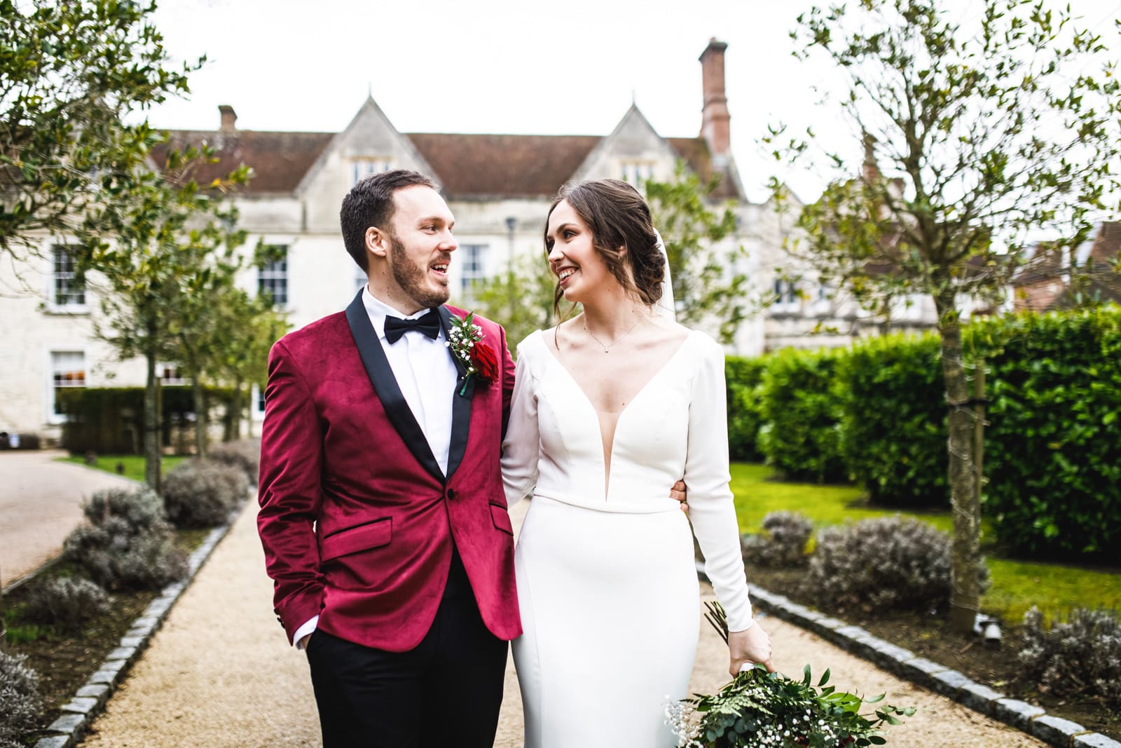 A bride in a white dress and a groom in a red velvet jacket walk hand in hand outside, smiling at each other, with greenery and the grand Froyle Park wedding venue behind them—captured perfectly by a Hampshire wedding photographer.